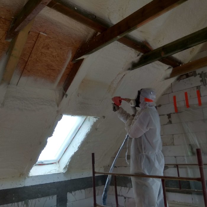 Pracownik w kombinezonie ochronnym wykonujący natrysk pianki poliuretanowej na poddaszu z widocznymi oknami dachowymi i drewnianą konstrukcją.